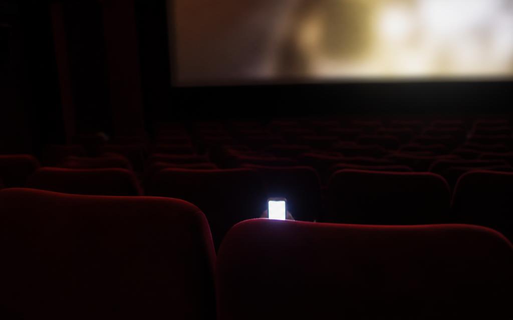 Bright phone screen glowing in a dark cinema row while other viewers watch the film